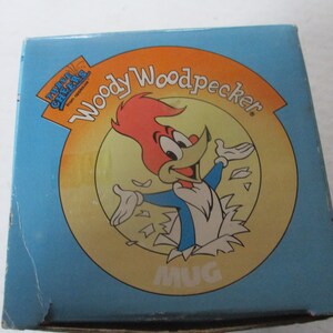 Puede incluir: Una caja azul y amarilla con una imagen de dibujos animados de Woody Woodpecker. La caja dice "Three Cheers from Topps" y "Woody Woodpecker" con una imagen de taza en el centro.