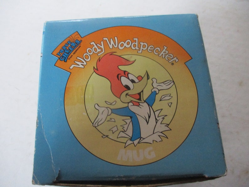 Puede incluir: Una caja azul y amarilla con una imagen de dibujos animados de Woody Woodpecker. La caja dice "Three Cheers from Topps" y "Woody Woodpecker" con una imagen de taza en el centro.