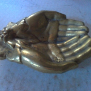 May include: A vintage brass sculpture depicting two open hands in a cupped position. The sculpture has a warm, golden hue and shows signs of age and wear. The hands are detailed, with visible fingers and a textured surface.