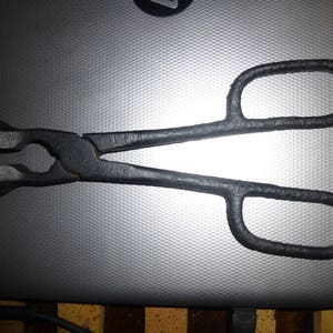 May include: Black metal tongs with looped handles and a scissor-like design. The tongs have a textured surface and are designed for gripping and manipulating objects. The tool is likely used in a workshop or for blacksmithing.