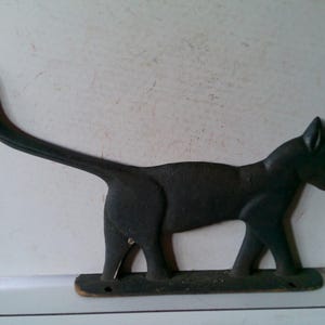May include: A black metal cat silhouette, likely a wall decoration or hook. The cat is depicted in profile with a long, curved tail and is mounted on a rectangular base. The cat's form is solid and the overall design is simple.