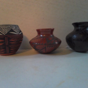 May include: Three small pottery vases with different designs. The vase on the left has a black and white geometric pattern. The middle vase has a red and black geometric pattern. The vase on the right is black.