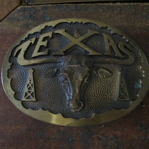 May include: A brass belt buckle with a raised design of a longhorn steer's head with horns, oil derricks, and the word "TEXAS" in raised letters.