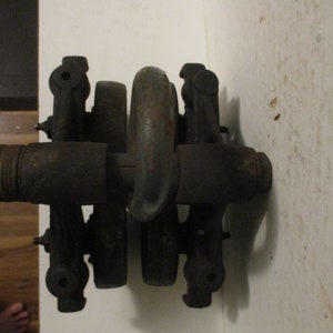 May include: A close-up of a dark, metallic mechanical component. The object has a series of cylindrical and curved parts, with a central ring-shaped element. The metal shows signs of age and wear, with a slightly rusted appearance. The component is mounted against a white wall.
