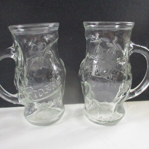 May include: Two clear glass mugs with handles. The mugs have a raised design of a bird and the text "Budweiser" on the front.