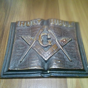 May include: A rectangular bronze-toned metal plaque shaped like an open book. The text HOLY BIBLE is embossed at the top. Centered on the pages is a raised Masonic square and compass symbol with a letter G in the middle and a brick pattern below.