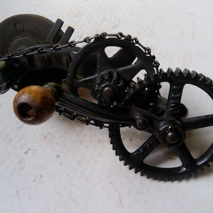 May include: An antique, cast iron sharpening tool with a wooden handle. The tool features a grinding wheel, gears, and a chain. The metal components are dark and show signs of age and use. The wooden handle is a light brown color.