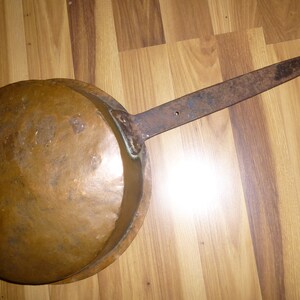 May include: A vintage copper pan with a long, rusty iron handle. The pan is round and has a slightly domed bottom. The handle is attached to the pan with a metal rivet.