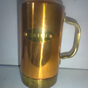 May include: A vintage copper-colored metal mug with a brass handle and base. The mug has a cylindrical shape with a slightly rounded top. The word "CHEESEO" is displayed on an oval-shaped brass plate on the front of the mug.