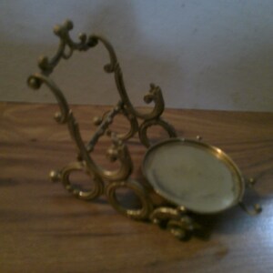 May include: A gold-toned metal stand with an ornate design. The stand has a round top and a decorative base.