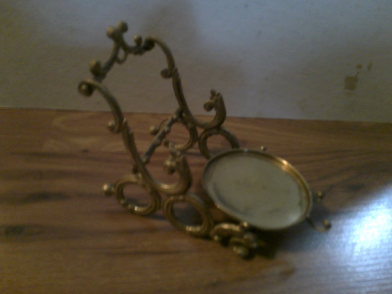 May include: A gold-toned metal stand with an ornate design. The stand has a round top and a decorative base.