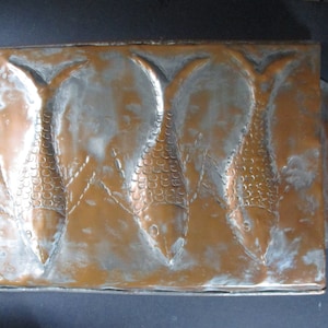 May include: A copper plaque with three fish embossed in the metal. The fish are facing to the right and have scales and fins. The plaque has a slightly aged patina.