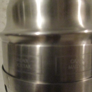 K&ouml;nnte beinhalten: Ein gl&auml;nzender Cocktailshaker aus Edelstahl. Der Shaker hat einen zylindrischen K&ouml;rper mit ausgestelltem Ober- und Unterteil. Die Texte "RIGHT MORTAR" und "CAJUN MACHINE" sind auf der Oberfl&auml;che eingraviert.