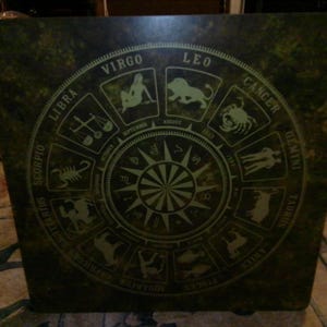 May include: A square, dark green table featuring a circular zodiac chart printed in a lighter green tone. The design displays all twelve astrological signs, including Libra, Virgo, Leo, Cancer, Gemini, Taurus, Aries, Pisces, Aquarius, Capricorn, Sagittarius, and Scorpio, arranged around a central sunburst pattern with corresponding months.
