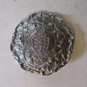 May include: Silver-toned metal belt buckle with an ornate design featuring a shield with three horizontal bars and a crown above it.