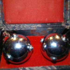 May include: Two shiny, silver metal balls with small protruding knobs, resting on a red velvet surface inside a decorative box. The box has a black exterior with a floral pattern and a red interior.