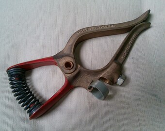 May include: A vintage welding clamp with a red handle and a black spring. The clamp is made of bronze-colored metal and has the text "Tweco Products Inc" on the top arm. A gray metal ring and a silver bolt are also visible.