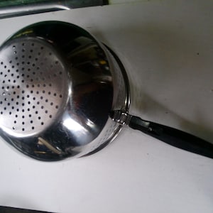 May include: A stainless steel colander with a black handle. The colander has numerous small holes for draining liquids. The handle is attached with a metal bracket.