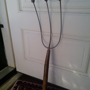 May include: A vintage, three-pronged garden tool with a wooden handle and metal tines. The handle is a dark brown color, and the tines have a rounded top. The tool is leaning against a white door.