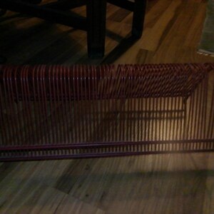 May include: A vintage red metal record holder. The holder has a rectangular shape with vertical bars and a woven top section. It is designed to hold vinyl records and is a retro storage solution.