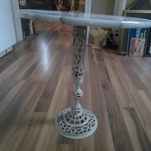 May include: A white, ornate side table with a round, clear top. The table features a decorative, openwork design on the base and central column. The table is approximately 28 inches tall.