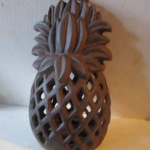 May include: A brown, decorative pineapple-shaped object with a textured surface and diamond-shaped cutouts. The top features detailed leaf-like elements, and a small loop is at the top for hanging. This item is likely a home decor piece.
