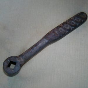 May include: A dark, weathered metal ratchet handle featuring a square drive socket at one end. The tool has a long, cylindrical shaft with a textured, worn surface and visible stamped lettering along the length of the handle, showing signs of age.