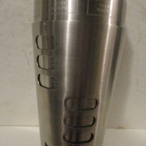K&ouml;nnte beinhalten: Ein Cocktailshaker aus Edelstahl mit den Worten "BRONX COCKTAIL" und "CAJUN MARTINI" auf der Oberfl&auml;che. Der Shaker hat eine konische Form mit mehreren l&auml;nglichen &Ouml;ffnungen und Messmarkierungen.
