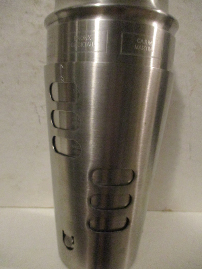 K&ouml;nnte beinhalten: Ein Cocktailshaker aus Edelstahl mit den Worten "BRONX COCKTAIL" und "CAJUN MARTINI" auf der Oberfl&auml;che. Der Shaker hat eine konische Form mit mehreren l&auml;nglichen &Ouml;ffnungen und Messmarkierungen.