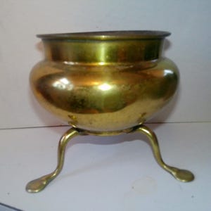 May include: A vintage brass pot with a rounded body and a flared rim. The pot is supported by three curved legs. The brass has a warm, golden hue and shows signs of age, adding to its antique charm. This decorative piece could be used as a planter or a decorative item.