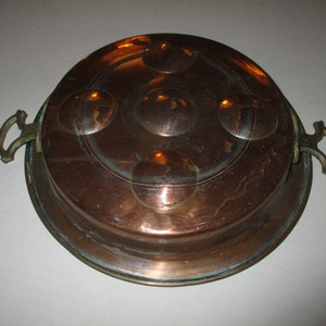 May include: A copper-toned, round serving dish with a lid. The lid features four raised circular designs and is flanked by two brass handles. The dish has a vintage appearance, suggesting it is an antique or a well-preserved piece.