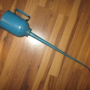 May include: A vintage blue oil can with a long, slender spout and a handle. The can is made of metal and has a slightly worn appearance, suggesting it has been used. The oil can is positioned on a wooden surface.