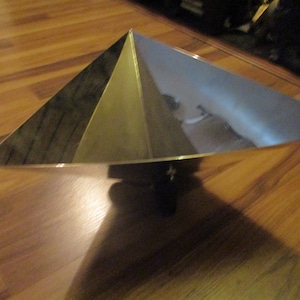 May include: A metallic pyramid-shaped decorative bowl with a reflective interior. The bowl has a triangular base and sides that slope upwards to a point. The bowl is sitting on a wooden surface.