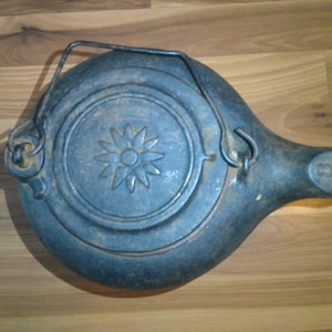 May include: A vintage cast iron kettle with a black finish and a flower design. The kettle has a handle and a spout.