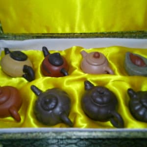 May include: A collection of eight small, brown and black ceramic teapots in a yellow box. The teapots are arranged in two rows of four, with each teapot having a different design.