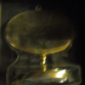 May include: A clear glass bottle stopper with a rounded top and a flared base. The stopper has a small, round knob at the top and a long, thin stem. The glass has a slight yellow tint.