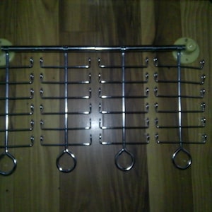May include: A silver-colored metal jewelry display rack with multiple hooks. The rack is mounted on a wooden surface with two suction cups. The hooks are arranged in rows, designed for hanging necklaces, bracelets, or other accessories.