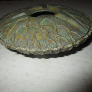 May include: A green and brown metal ashtray with a decorative design. The ashtray has a circular shape with a center hole and a scalloped edge.