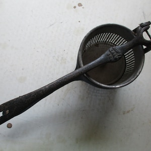 May include: An antique, cast-iron food mill with a perforated bowl, a pressing disc, and a long handle. The handle has a loop at the end for hanging. The food mill is dark gray and appears to be well-used.
