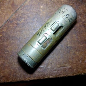 May include: A vintage brass instrument with the word "MAJOR" engraved on its side. The cylindrical tool has two rectangular cutouts and a ridged end. The top of the instrument has engraved markings, including "b.B.C.C."