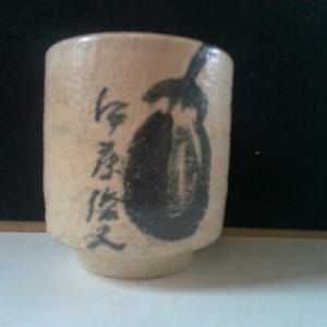 May include: A beige ceramic teacup with a black and white illustration of an eggplant and Japanese characters.