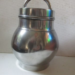 May include: A shiny, silver-colored metal container with a rounded body and a curved handle. The container has a lid and a base, and it reflects light, creating highlights and shadows.