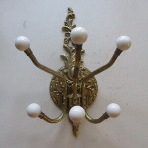 May include: Ornate brass wall-mounted coat rack with five curved arms, each tipped with a white ceramic ball. The rack features a decorative floral and scroll design on the central mounting plate.