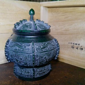 May include: A dark green, lidded jar with intricate carved designs. The jar has a rounded body and a pedestal base. The lid is topped with a small, rounded finial. The jar is placed in front of a wooden box.