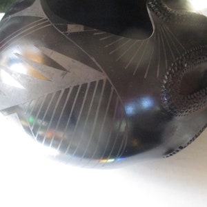 May include: Black ceramic bowl with a geometric design. The bowl has a wide, rounded opening and a narrow base. The design is made up of lines and curves that create a pattern of triangles and squares.