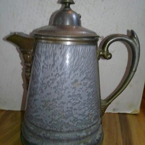 May include: Antique silver and gray speckled coffee pot with a lid and handle. The pot has a spout and decorative brass accents. The pot is likely made of metal and is designed for serving hot beverages.