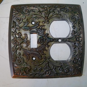 May include: Ornate, square electrical outlet cover with a switch and two outlets. The cover is bronze-colored with a floral and leaf design. The design is raised, creating a textured appearance.