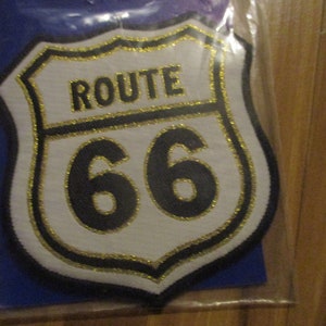 May include: A white, gold, and black Route 66 patch with a gold glitter outline. The patch is shaped like a shield and has the text "ROUTE 66" in black letters with a gold glitter outline.