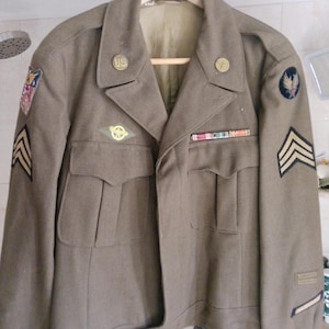 May include: An olive drab military jacket featuring a notched collar, two front pockets, and various patches and pins. The jacket includes a US collar pin, a ribbon bar, chevron sleeve patches, and shoulder insignia, all displayed on a wooden hanger.
