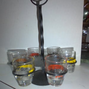 May include: A black metal shot glass holder with six clear shot glasses. The shot glasses have colorful designs on them.
